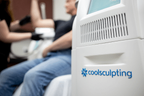 A person undergoing a CoolSculpting treatment in a clinic. The image focuses on the CoolSculpting machine in the foreground, with a healthcare professional administering the treatment to the patient's arm in the background. The CoolSculpting logo is visible on the machine.