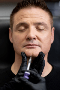 A man is receiving a cosmetic injection in his chin area while sitting on a black chair. The procedure is being performed by someone wearing black gloves. The man has his eyes closed and appears relaxed.