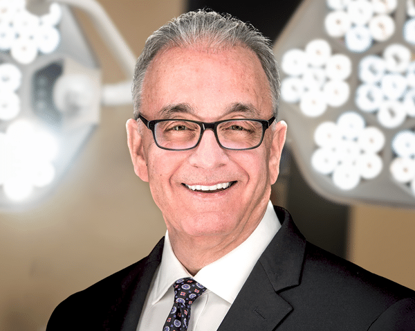 A man with graying hair and glasses is smiling. He is wearing a dark suit, white shirt, and a patterned tie. The background includes bright circular lights.