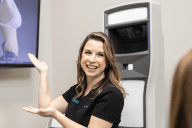 A woman with long brown hair is smiling and gesturing with both hands in front of a large, modern-looking machine. She is wearing a black shirt and standing indoors.