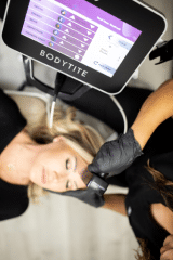 A person is lying down while another person, wearing black gloves, uses a device on their forehead. The device's screen, labeled "BODYTITE," displays various icons and text. The scene appears to be in a clinical or spa setting, suggesting a cosmetic or medical treatment is being performed.