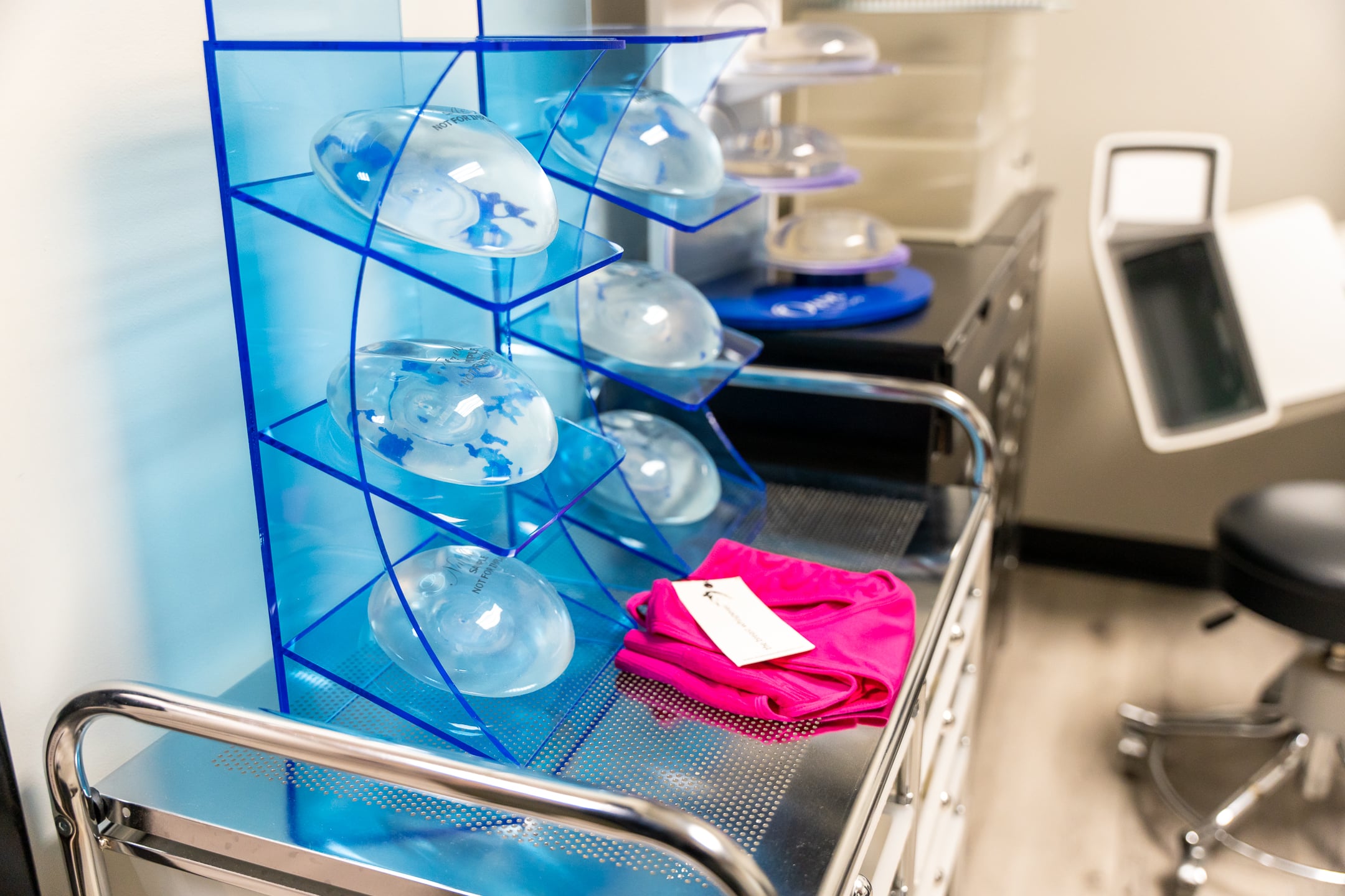A medical room with various clear silicone breast implants displayed on blue stands on a metal table. There are also bright pink fabric pieces with a tag placed on the table. In the background, there is a chair, a small electronic machine, and organized shelves.