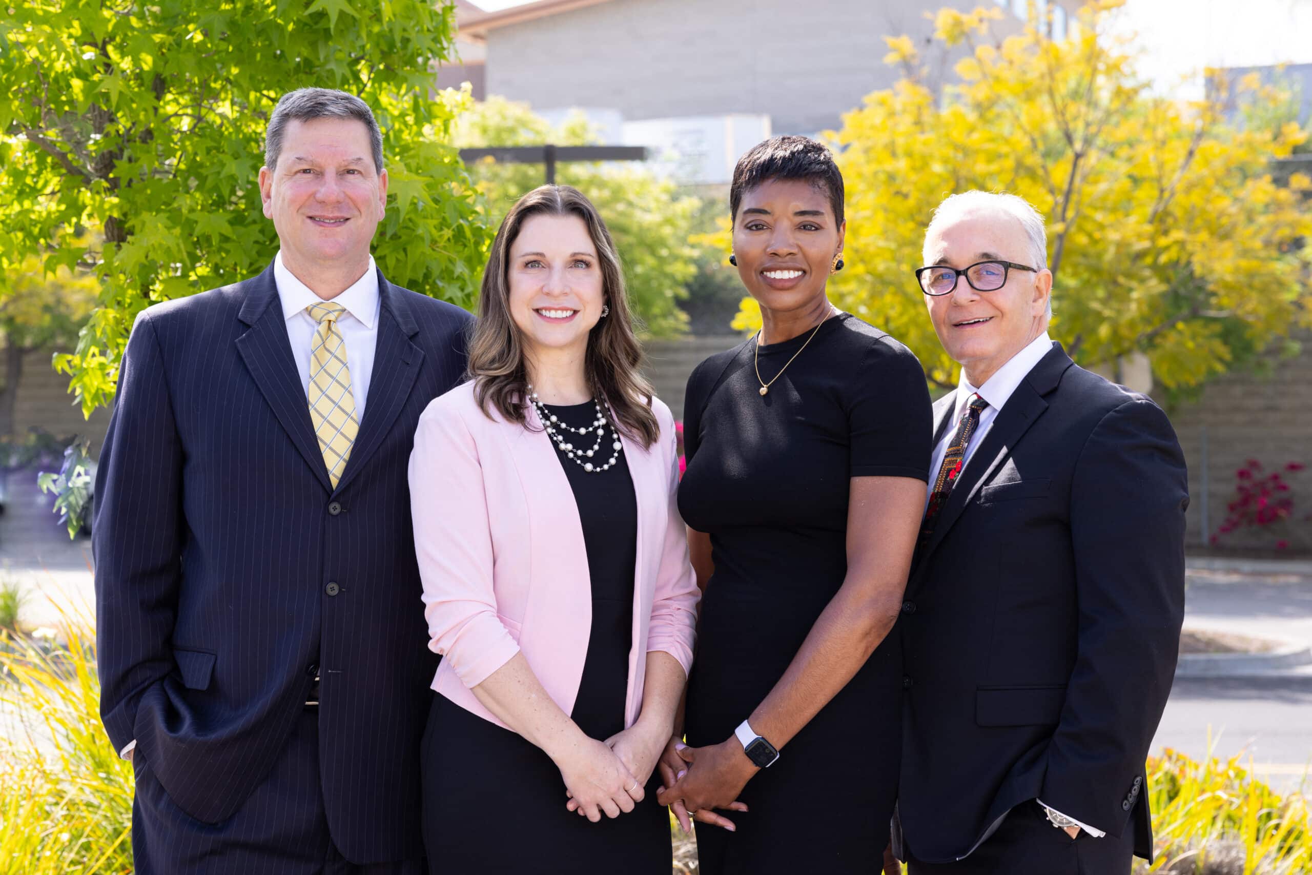 Four professionally dressed individuals standing outdoors, smiling, and posing for a group photo. They are in front of green trees and a building is visible in the background. All are wearing business attire: two men in suits and two women in dresses, one of whom is wearing a light pink blazer.