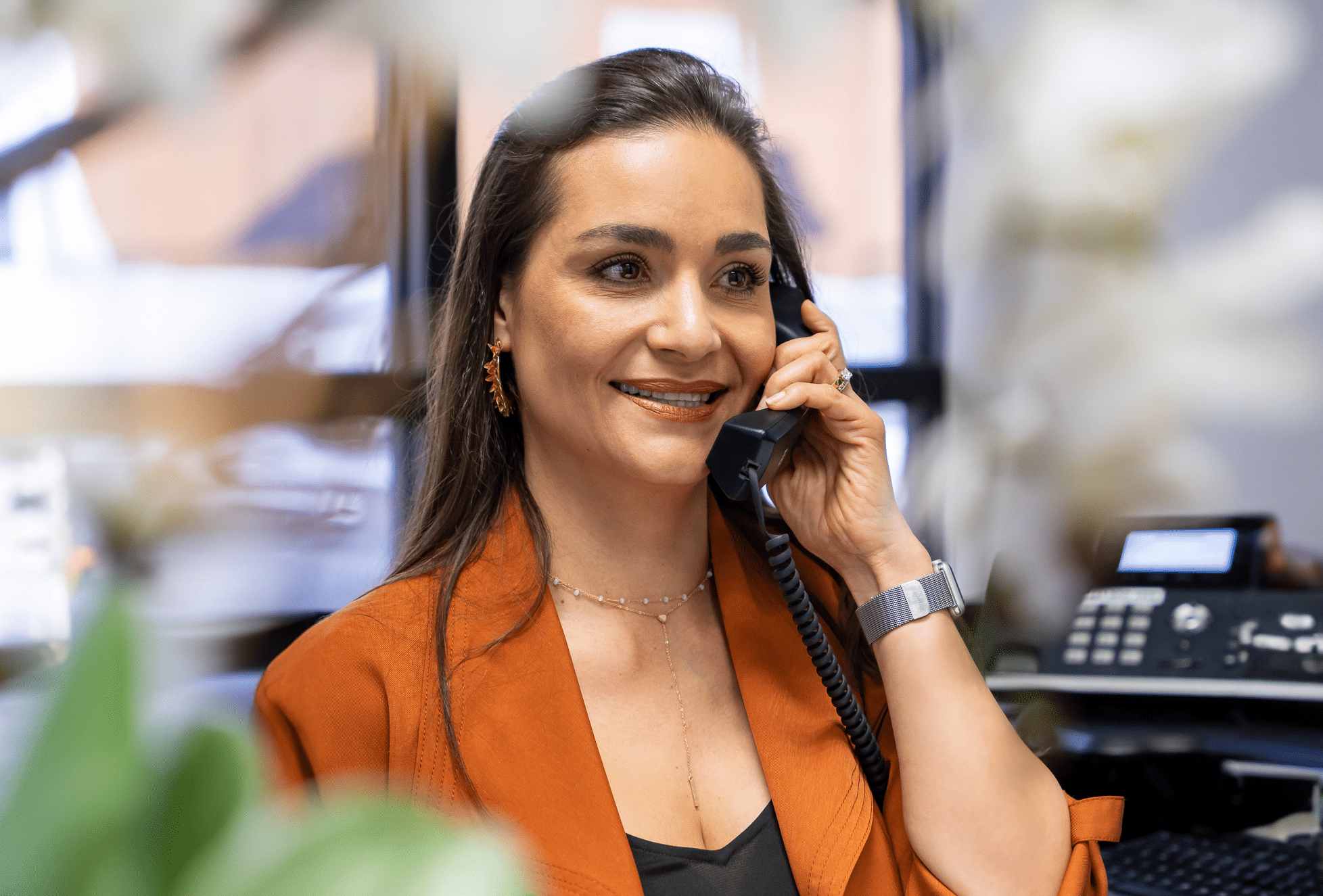 A woman with long hair, wearing an orange blazer and a black top, is smiling while talking on a phone in an office setting. In the foreground, there are some blurred white flowers, and in the background, an office phone and other office equipment are visible.
