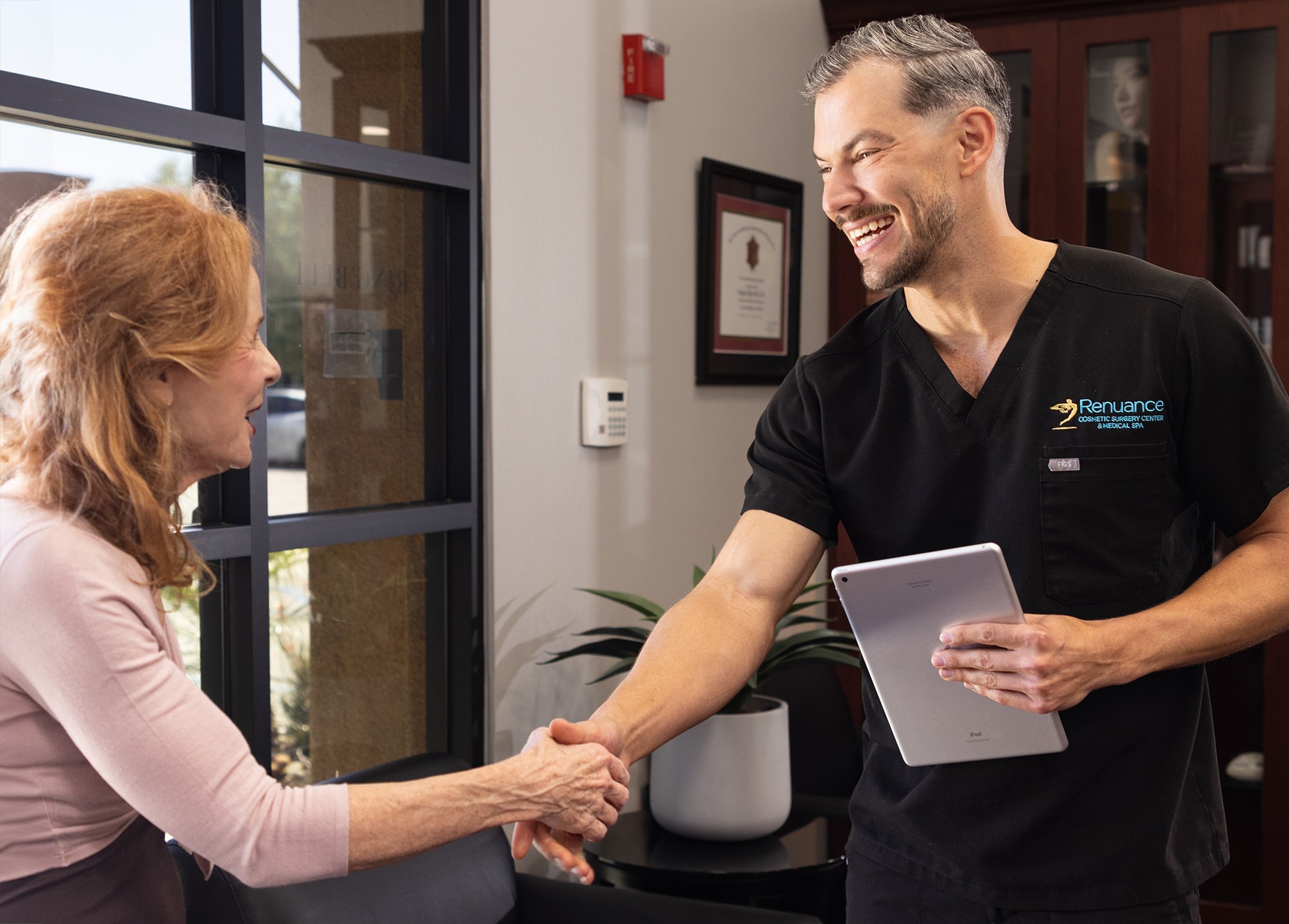 A doctor, wearing a black uniform with the logo "Renuance Cosmetic Surgery Center," shakes hands with a woman in a waiting room. The doctor holds a tablet in his left hand and is smiling. The woman has red hair and is wearing a light pink sweater. The background includes a window, a small plant, and framed certificates on the wall.