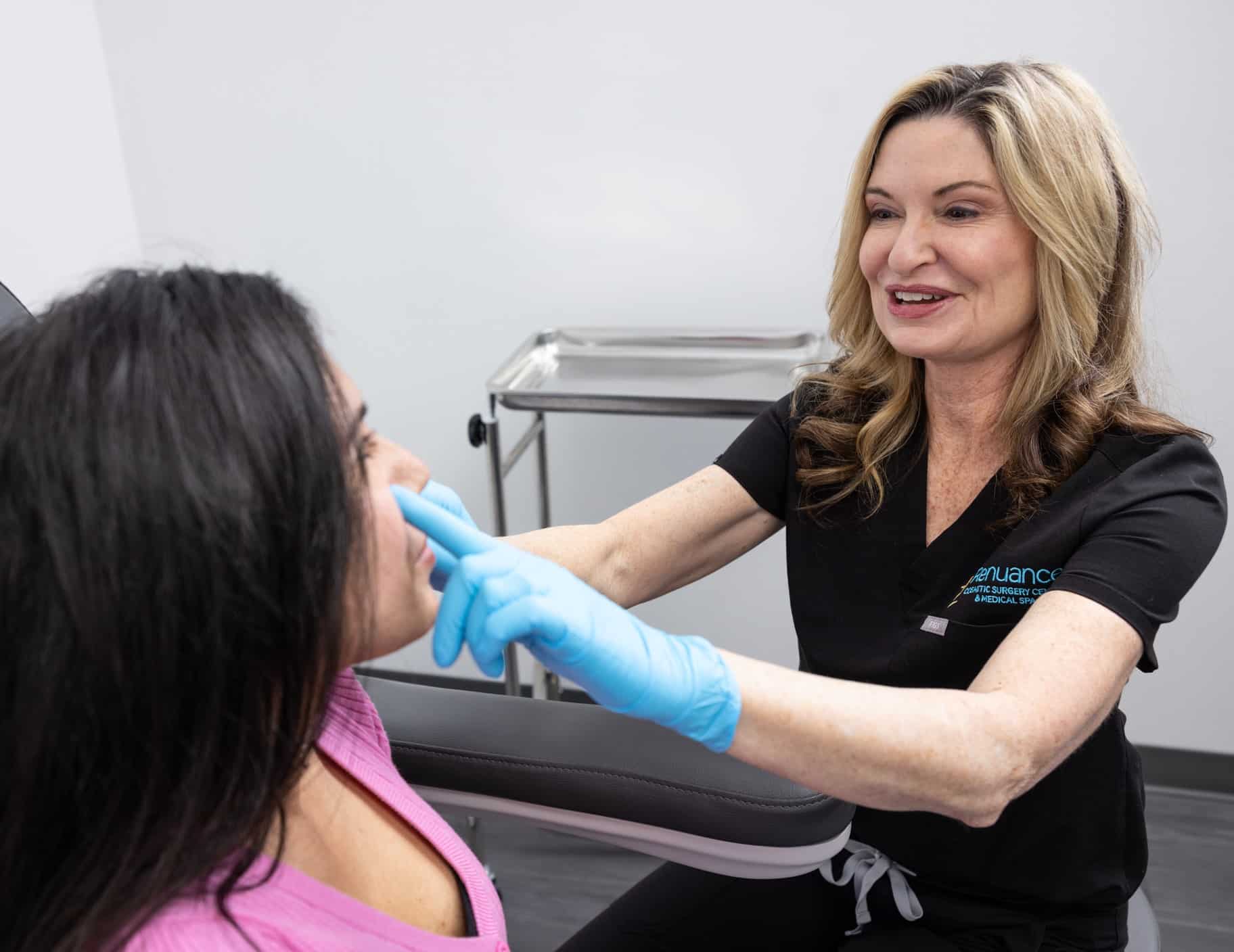 A healthcare professional wearing black scrubs and blue gloves is examining a patient's face. The patient, who has long dark hair and is wearing a pink top, is seated in a chair. The professional is gently touching the patient's nose while smiling and talking to her. In the background, there is a medical tray on a stand. The setting appears to be a medical or cosmetic consultation room.