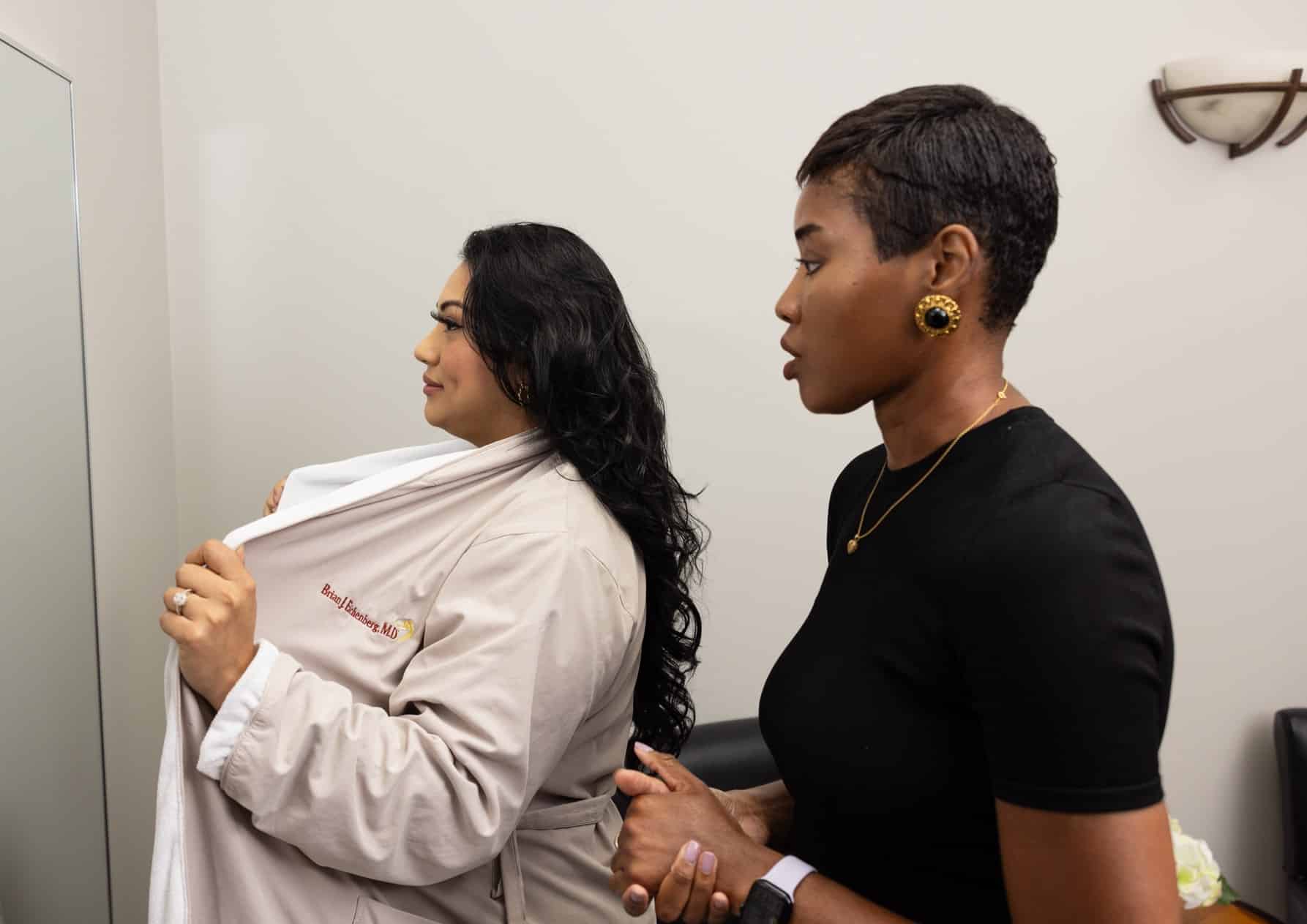 A woman in a black shirt is standing next to another woman who is wearing a beige coat and looking into a mirror. The woman in the coat has one hand on her chest, holding the coat slightly open. They appear to be in a professional setting, possibly a medical clinic or office.