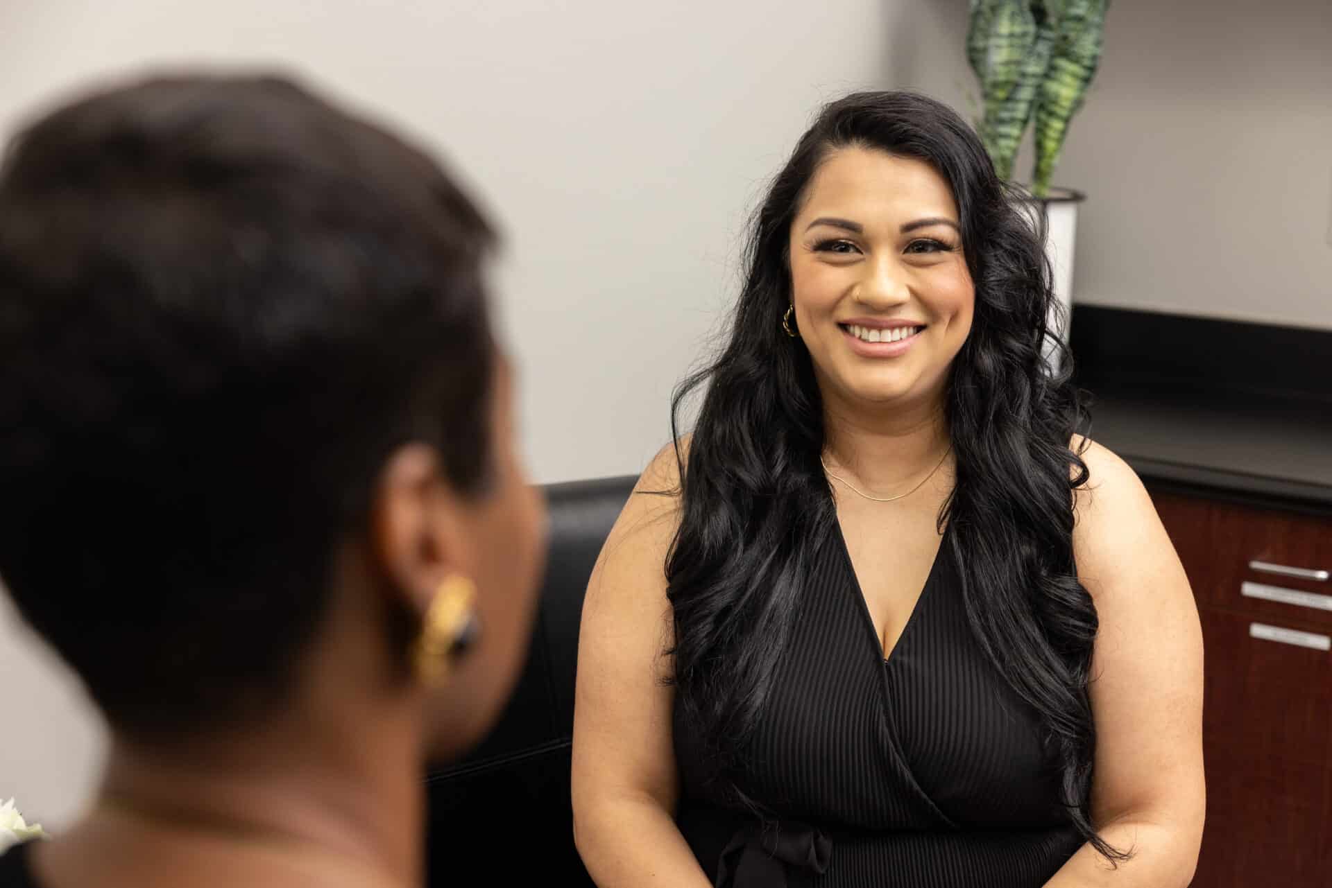 A woman with long dark hair smiles warmly at another person during a conversation in an office setting. The woman is dressed in a black top, while the other person's back is turned to the camera, revealing short hair and gold earrings. A plant and office furniture are visible in the background.