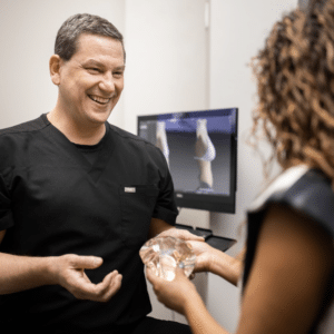 A man in black medical scrubs is smiling and holding a clear anatomical model, demonstrating something to a woman with curly hair. In the background, a computer screen displays a 3D model, possibly related to a medical procedure or anatomy. The setting appears to be a medical office or consultation room.