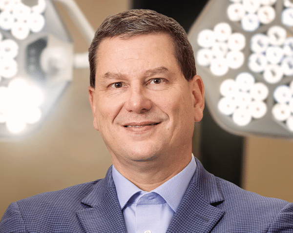 A man in a blue checkered blazer and light blue dress shirt is smiling at the camera. He has short dark hair and is standing in a well-lit environment, possibly an office or a professional setting, with round lights in the background.