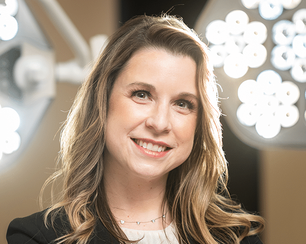 A woman with long blonde hair is smiling at the camera. She is wearing a black blazer over a light-colored shirt. The background features bright, circular lights, creating an illuminated effect around her head.