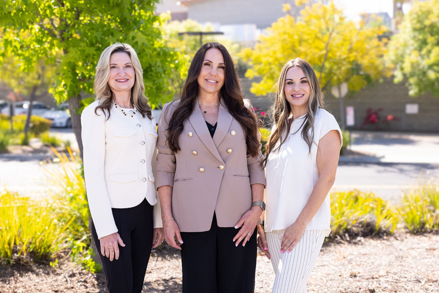 Three women are standing outdoors, posing for a photo. The woman on the left has blonde hair and is wearing a white jacket over a black top and dark pants. The woman in the center has long dark hair and is wearing a beige blazer with gold buttons and black pants. The woman on the right has long blonde hair and is wearing a white top and white pants. They are all smiling, and the background includes green trees and sunlight.