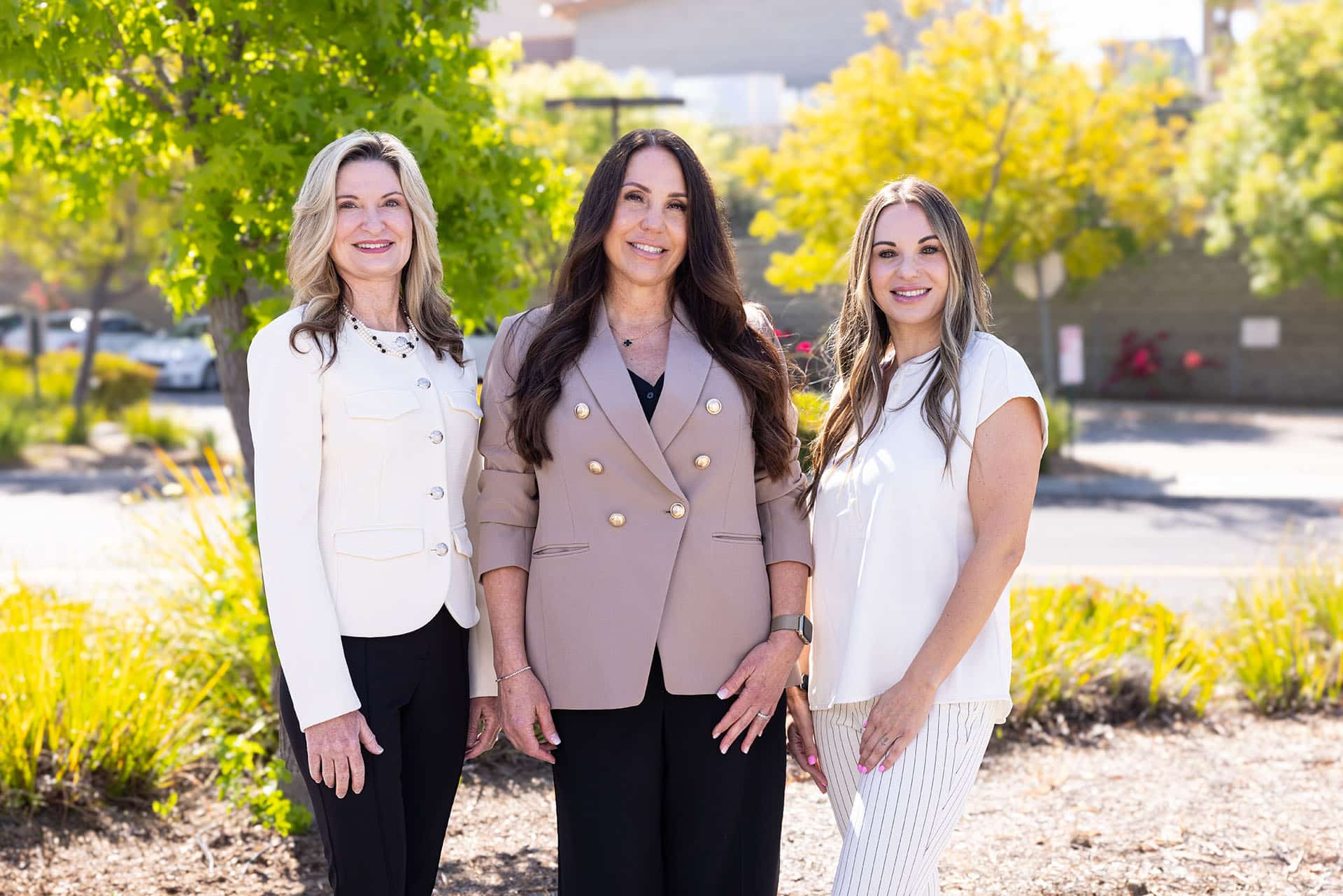 Three women are standing outdoors, posing for a photo. The woman on the left has blonde hair and is wearing a white jacket over a black top and dark pants. The woman in the center has long dark hair and is wearing a beige blazer with gold buttons and black pants. The woman on the right has long blonde hair and is wearing a white top and white pants. They are all smiling, and the background includes green trees and sunlight.