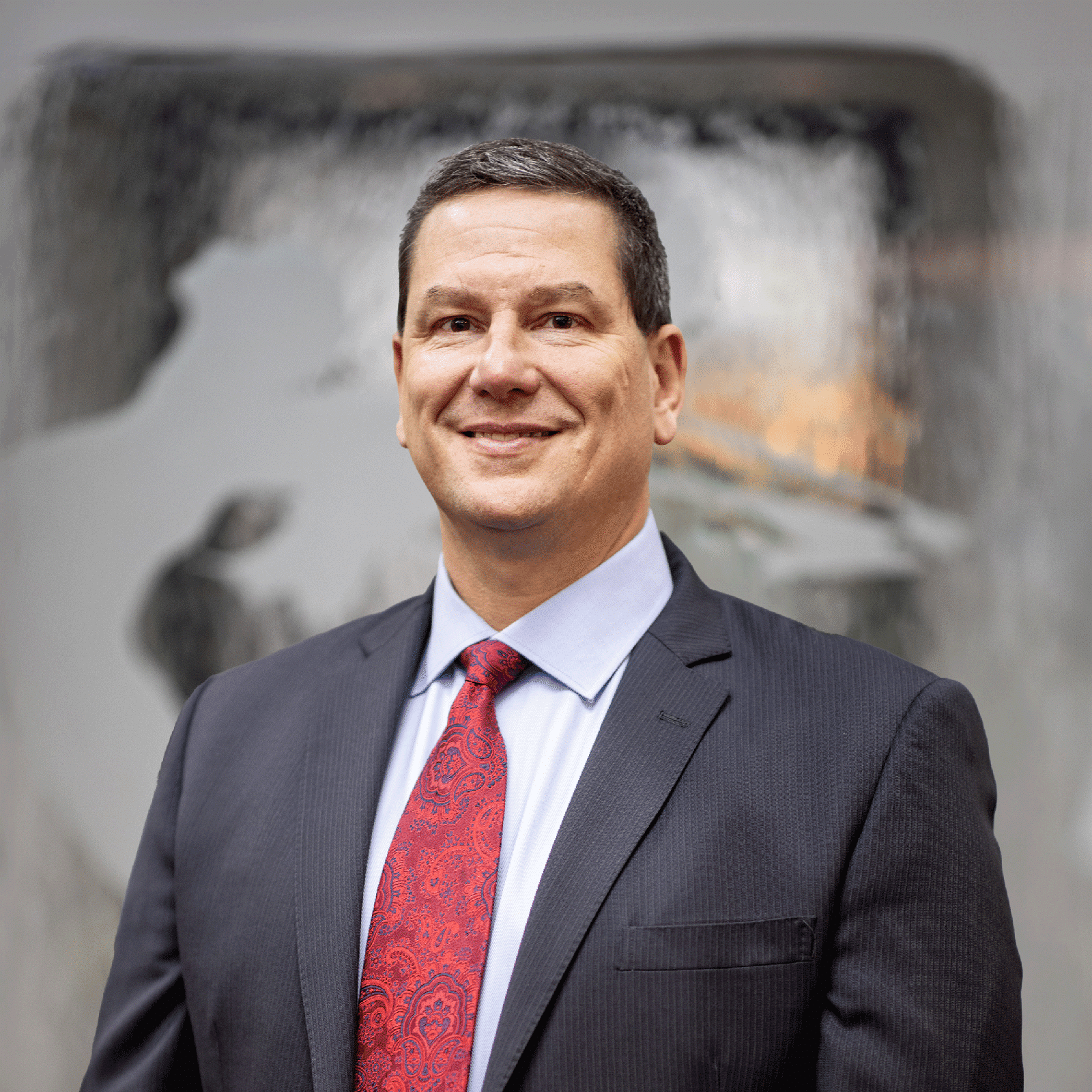 A man in a dark suit with a light blue dress shirt and a red tie stands smiling in front of a blurred background. He has short dark hair and appears to be in a professional setting.