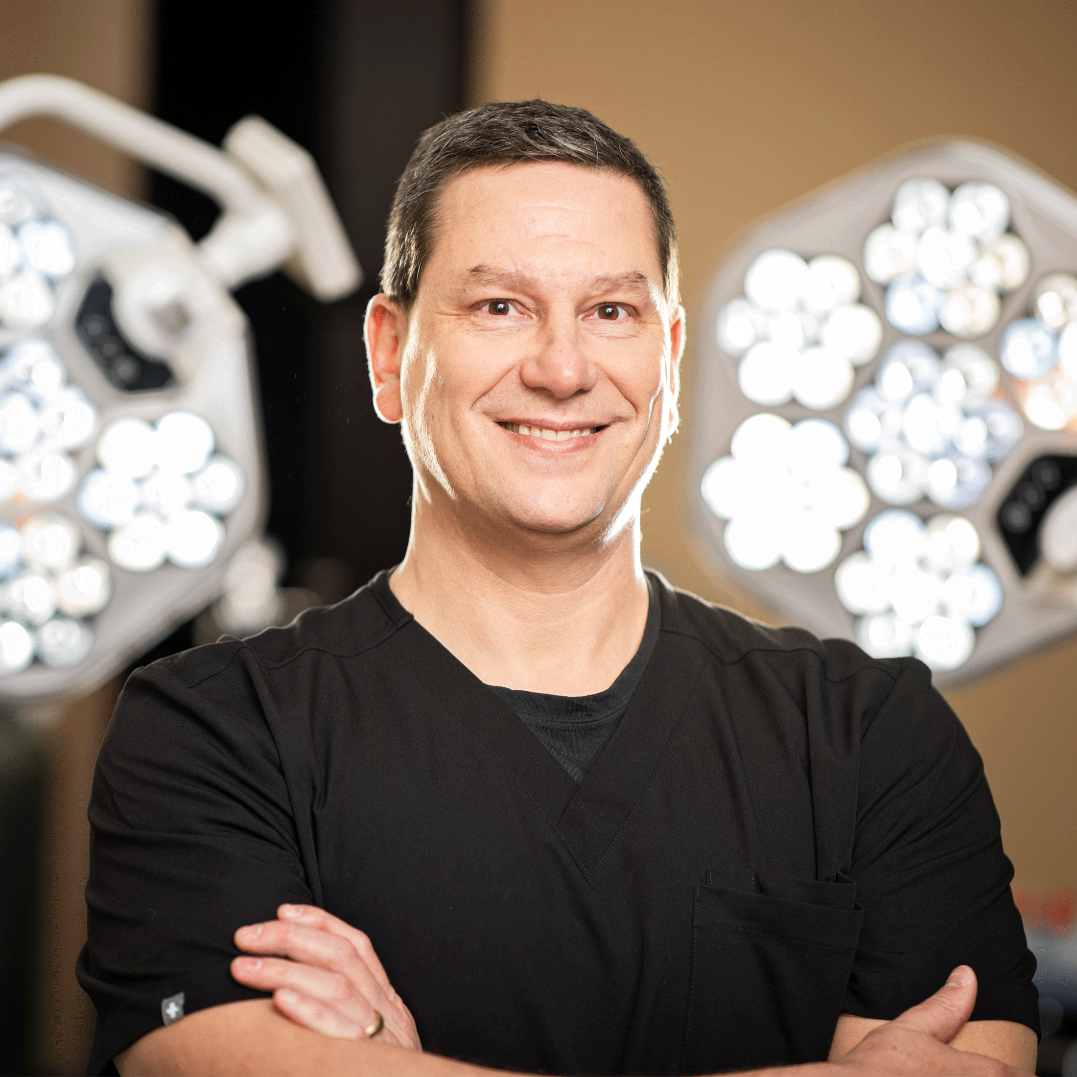 A man in medical scrubs stands with his arms crossed, smiling at the camera. He is in a room with bright surgical lights in the background.
