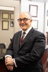 A man wearing glasses and a black suit with a patterned tie is smiling and standing in an office setting. He has short, gray hair and is leaning on a counter with framed certificates on the wall behind him.