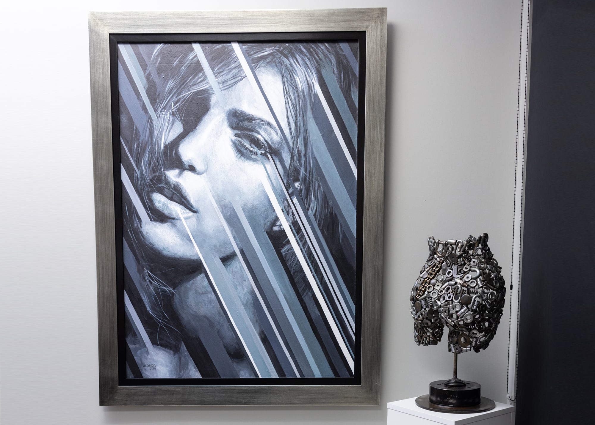 A monochromatic painting of a woman's face with slanted lines covering parts of the image is hung on a wall. Next to the painting, on a white pedestal, is a metallic sculpture with intricate, abstract detailing. The frame of the painting is a silver metallic color. The background wall is divided into white and dark gray sections.