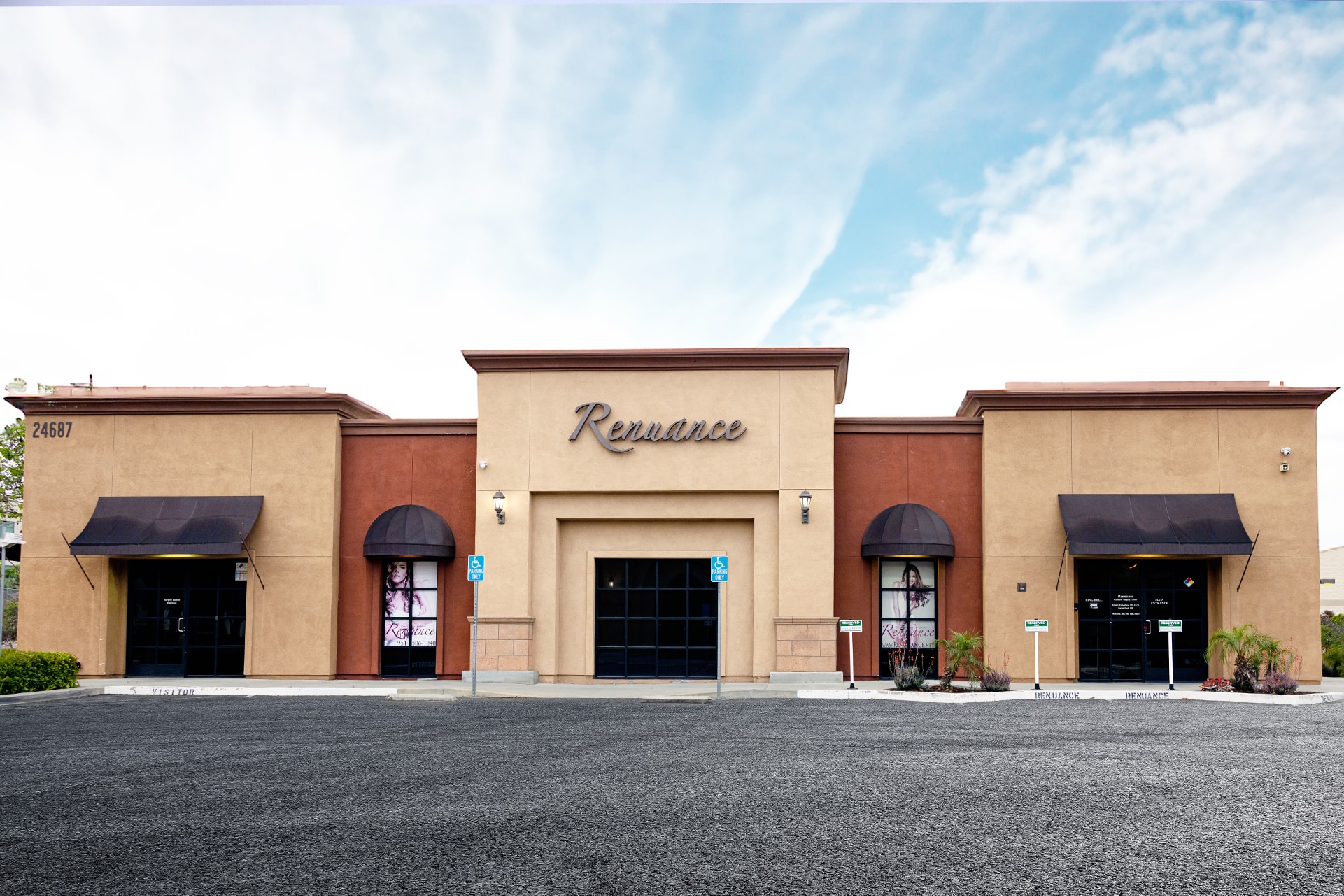 A single-story commercial building with three front entrances. The building features a blend of tan and reddish-brown exterior walls, with each entrance flanked by black awnings. The central entrance is more prominent with the word "Renaissance" displayed above it. There are accessible parking spaces and small landscaped areas with plants in front of the building. The sky is partly cloudy.