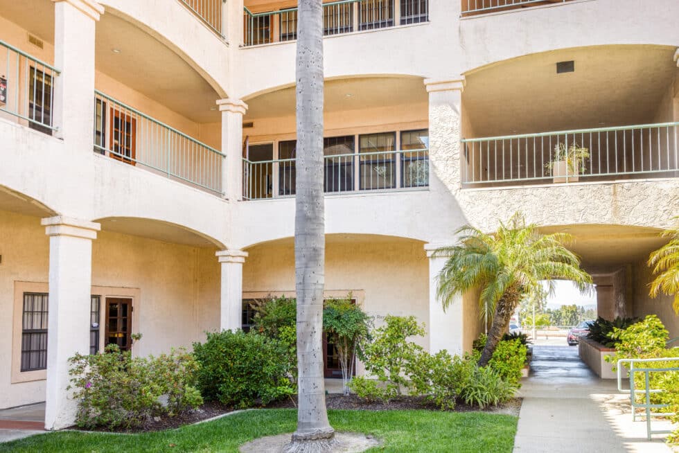 A two-story building with light-colored stucco walls and multiple archways. The upper level has balconies with green railings, and there are plants and shrubs along the ground level. A tall palm tree stands in front of the building, and there is a small walkway leading to an open area.