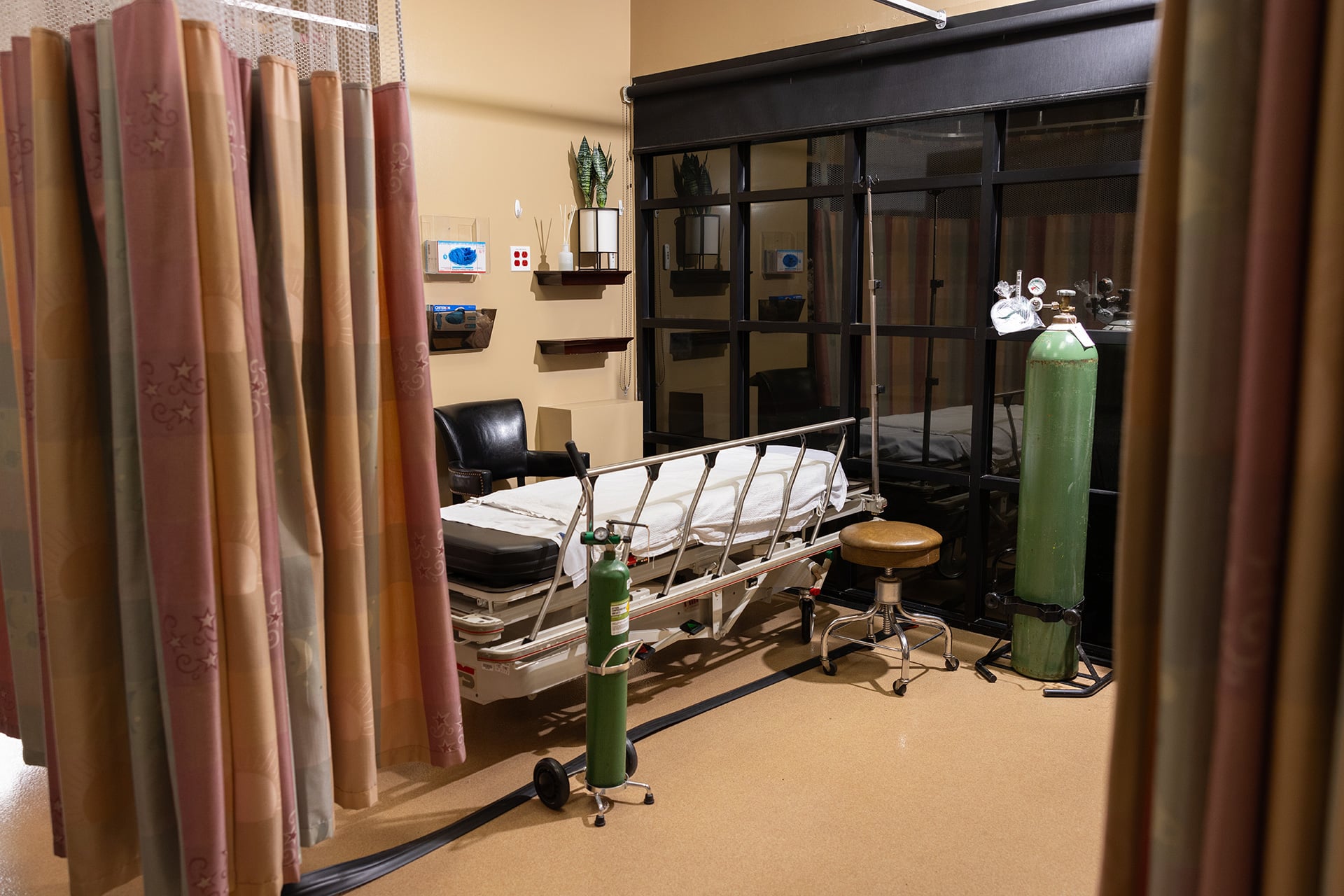 A hospital room with a bed, medical equipment, and privacy curtains. The bed has white linens and is equipped with guard rails. Next to the bed, there is a green oxygen tank. To the right of the bed, there is a brown stool. The room has beige walls and the privacy curtains are pink with patterns. On the far wall, there are shelves with various items and medical monitors.