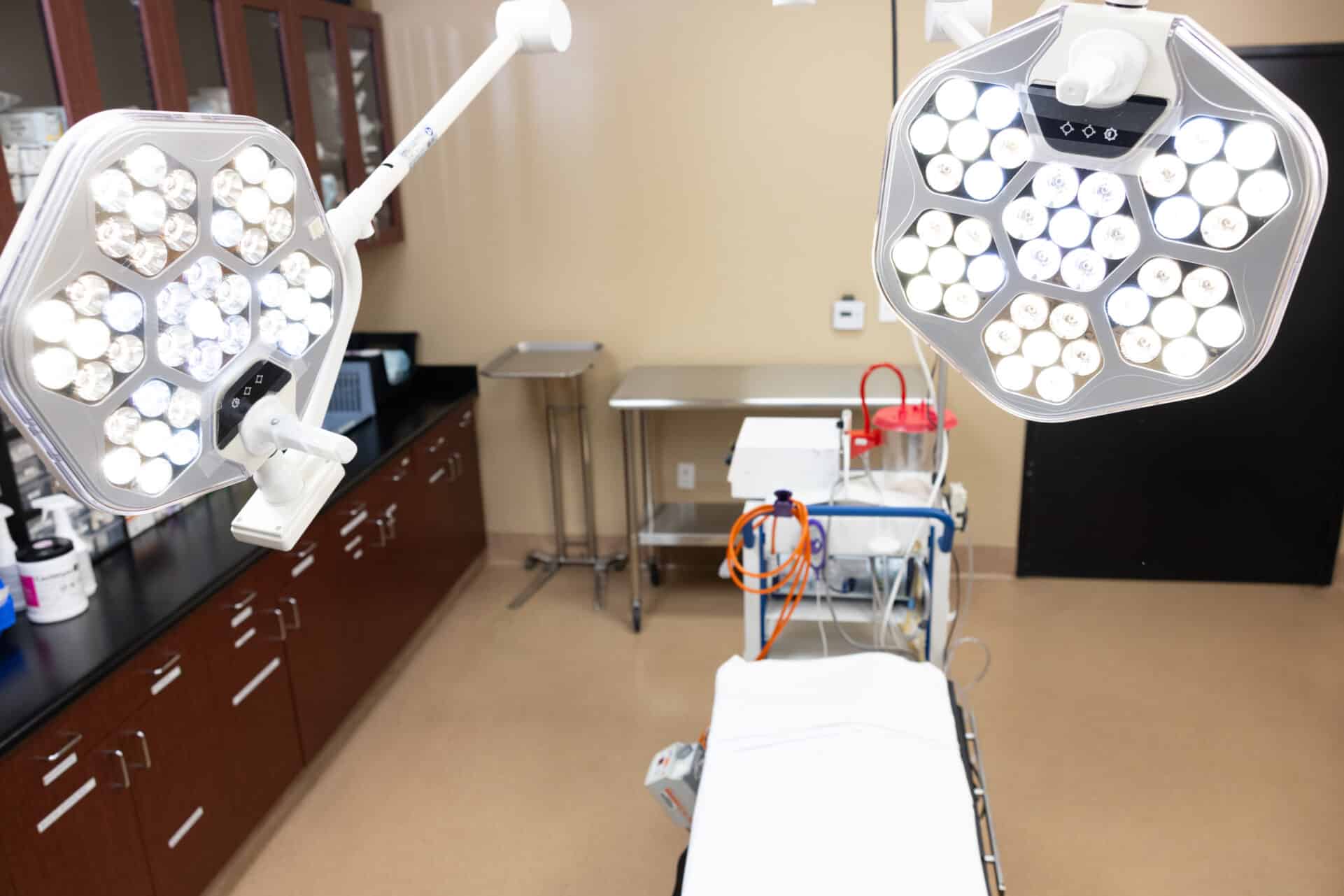 A medical examination room is equipped with bright overhead surgical lights, a patient examination table covered with a white sheet, and medical instruments on a stainless steel table. Cabinets and countertops with various supplies are visible along the walls. The room appears clean and well-organized.