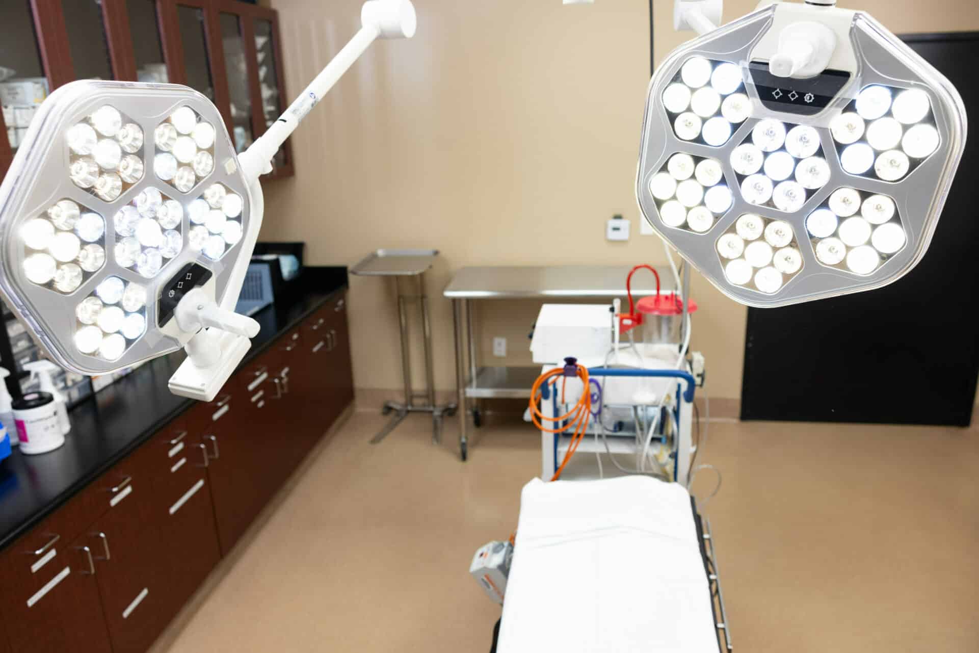 A medical examination room with overhead surgical lights, an examination table, and various medical cabinets and equipment. The room has bright, clean lighting and is equipped with modern medical tools.