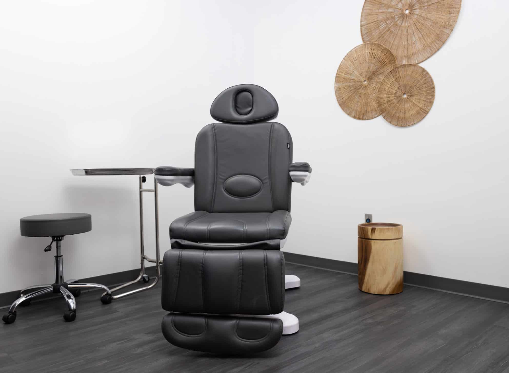 An empty medical examination room featuring a black leather recliner chair in the center. A small, round, black stool is positioned next to the chair on the left, and a metal tray table is adjacent to it. The room has white walls and dark, wooden flooring. Three decorative, circular, wicker wall hangings are displayed on the right side of the wall.