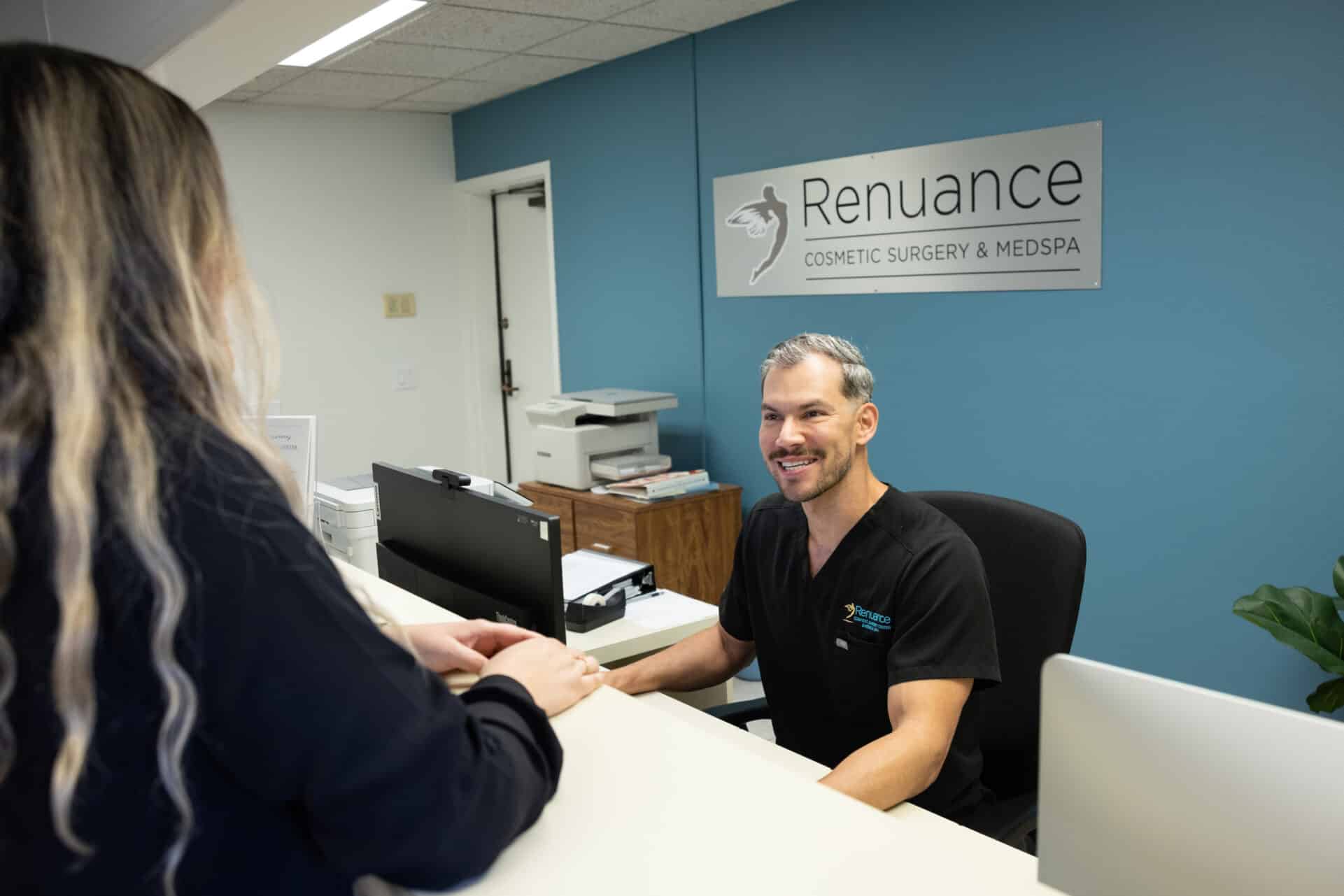 A medical professional in black scrubs, seated at a reception desk, smiles while speaking with a person standing on the other side of the desk. The background features a blue wall with a sign that says "Renuance Cosmetic Surgery & Medspa." The environment appears to be a welcoming medical office or clinic setting.