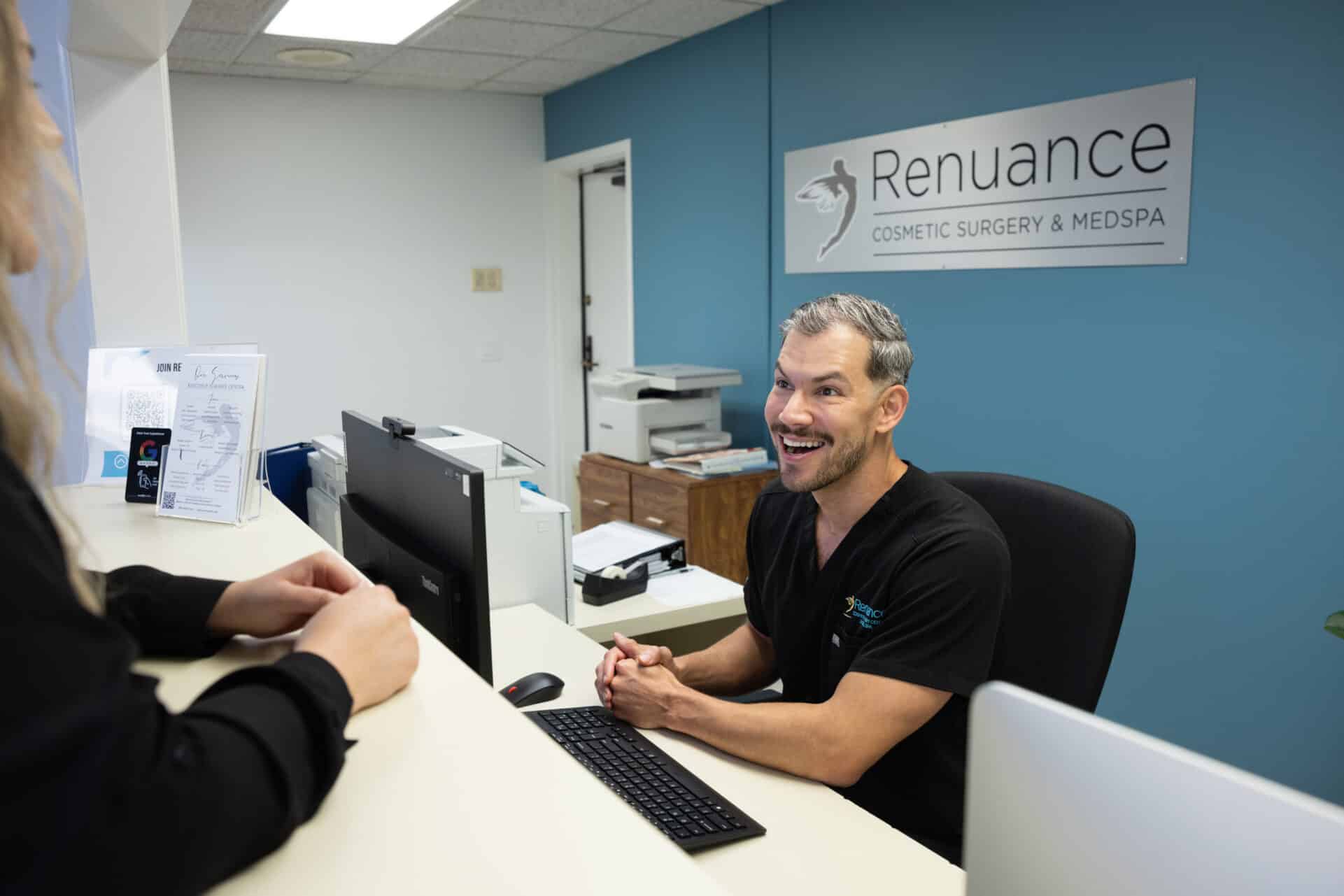 A person with short hair and facial hair is seated at a reception desk, smiling and speaking to another individual who is partially visible in the foreground. The reception area has a blue and white color scheme, with a sign on the wall that reads "Renuance Cosmetic Surgery & Medspa." A computer, keyboard, and documents are visible on the desk.