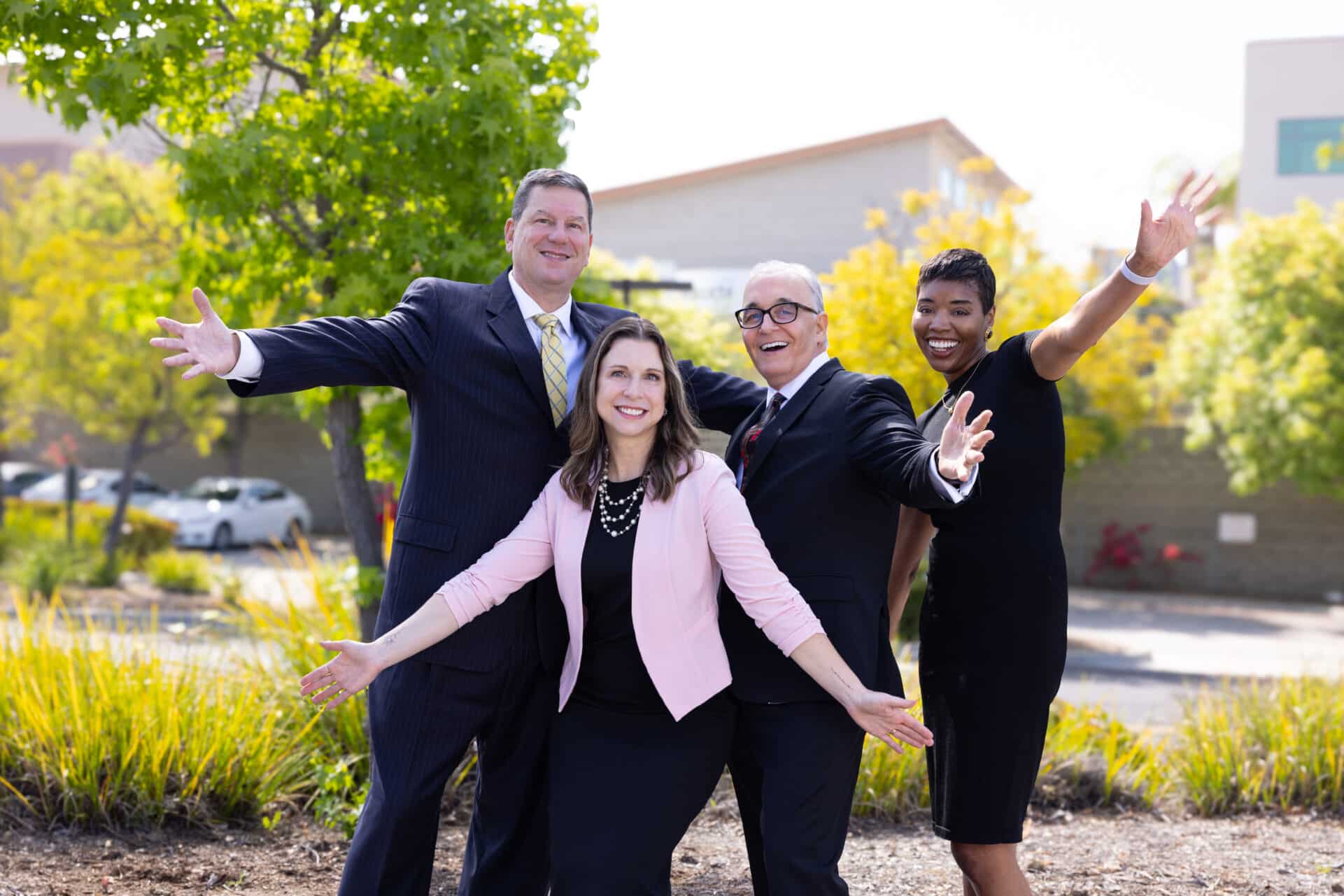 A group of four professionally dressed people are standing outdoors, smiling, and holding their arms out in a joyful gesture. There is a man in a suit and tie on the left, a woman in a pink blazer in the center, a man in a black suit and tie next to her, and a woman in a black dress on the right. They are standing in front of some greenery and a building. The atmosphere appears to be cheerful and energetic.