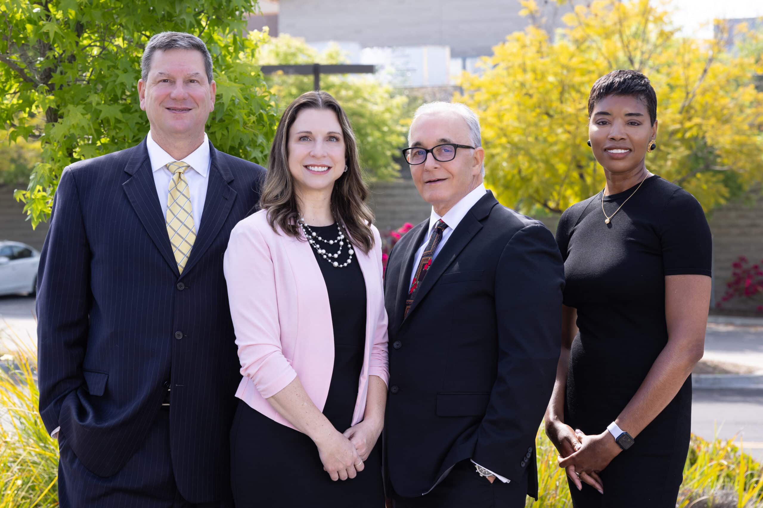 A group of four professionally dressed individuals standing outdoors. The first person on the left is a man in a dark suit and yellow tie. Next to him is a woman in a light pink blazer and black dress. To her right is an older man wearing glasses, a black suit, and a red patterned tie. The person on the far right is a woman in a black dress. They are all smiling and posing in front of a background with greenery and trees.