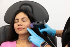 A woman sitting in a chair with her eyes closed while a medical professional wearing blue gloves uses a handheld device on the side of her face. The woman has dark hair and is wearing a pink top and a beaded choker necklace. The environment appears to be a clinical or medical setting.