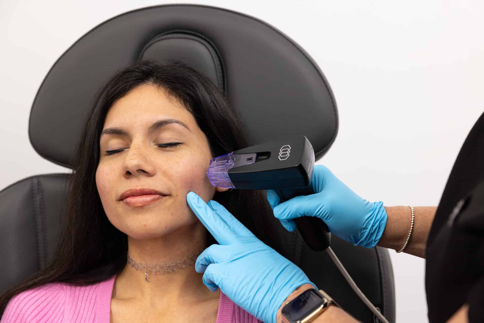A woman sitting in a chair with her eyes closed while a medical professional wearing blue gloves uses a handheld device on the side of her face. The woman has dark hair and is wearing a pink top and a beaded choker necklace. The environment appears to be a clinical or medical setting.