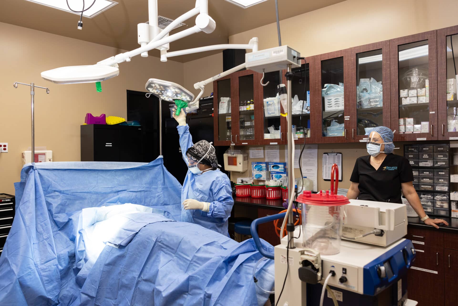 A medical team is performing surgery in an operating room. The surgeon, dressed in a blue surgical gown and gloves, is adjusting an overhead light while standing next to the patient, who is covered with a blue surgical drape. Another medical staff member, wearing scrubs, a hair cover, and a mask, is standing by a counter that has medical supplies and equipment. The room is well-lit and equipped with various medical instruments and storage cabinets.