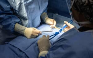 Two surgeons performing a surgery in an operating room. They are dressed in blue surgical gowns, gloves, and protective headgear, with their focus on the patient covered by a blue sterile drape. The surgeons are handling surgical instruments under bright surgical lighting.