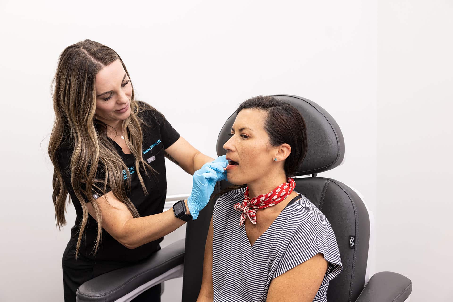 A healthcare professional wearing gloves is examining a patient's mouth. The patient, who is sitting in a medical chair, is wearing a red and white patterned scarf and a striped shirt. The setting appears to be a medical or dental office with a bright, clean environment.