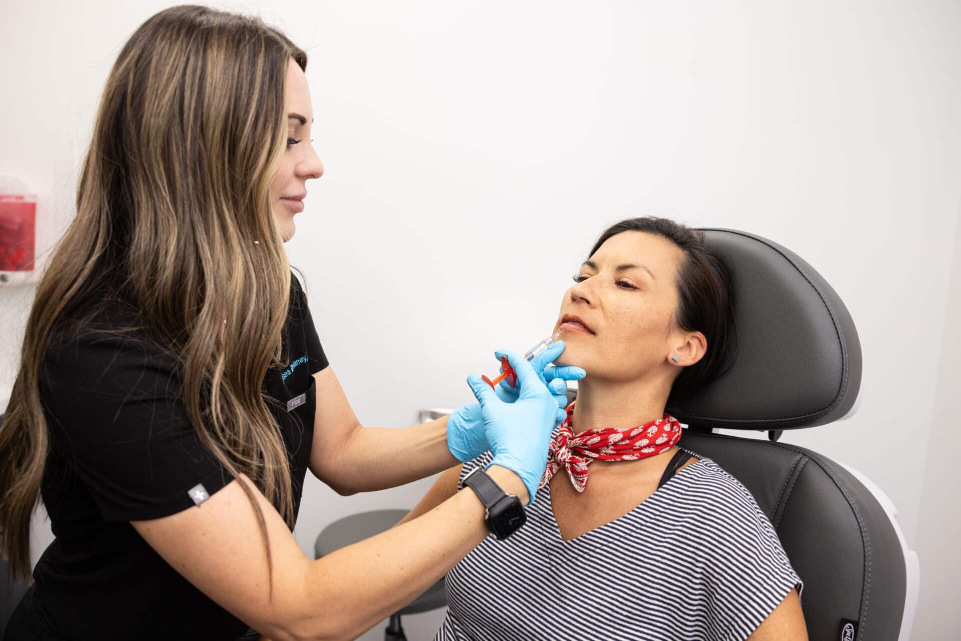 A woman with long brown hair is administering an injection to another woman seated in a chair. The seated woman is wearing a striped shirt and a red scarf around her neck. The healthcare professional is wearing blue gloves and a black uniform. The setting appears to be a medical or clinic room.