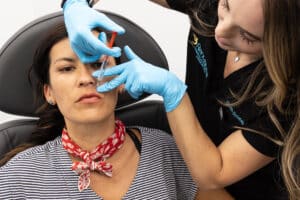 A woman is receiving a cosmetic injection in her face from a healthcare professional. The patient is lying back in a medical chair wearing a striped shirt and a red bandana around her neck. The healthcare professional, wearing blue gloves and a black uniform, is carefully administering the injection while holding the patient's face steady. Both are focused on the procedure.