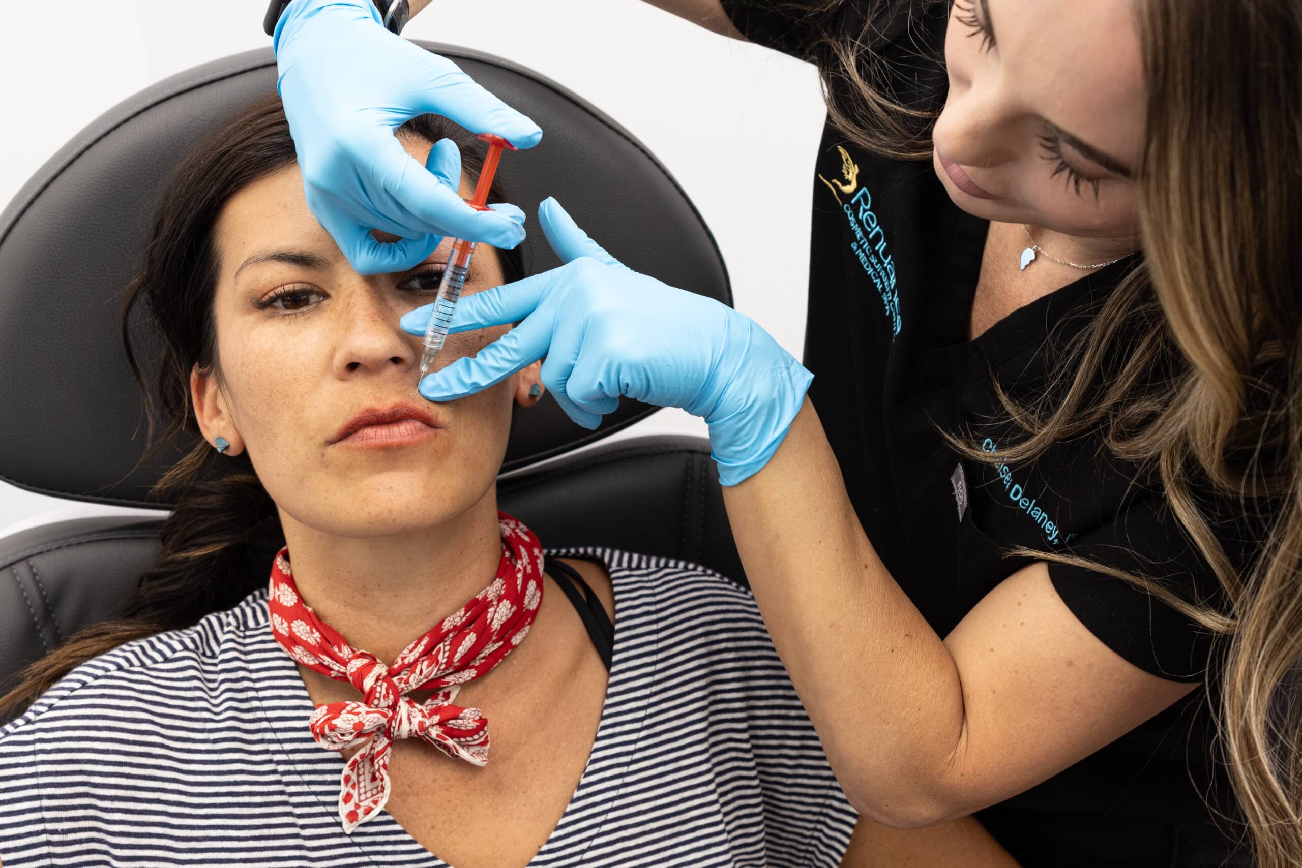 A woman is receiving a cosmetic injection in her face from a healthcare professional. The patient is lying back in a medical chair wearing a striped shirt and a red bandana around her neck. The healthcare professional, wearing blue gloves and a black uniform, is carefully administering the injection while holding the patient's face steady. Both are focused on the procedure.