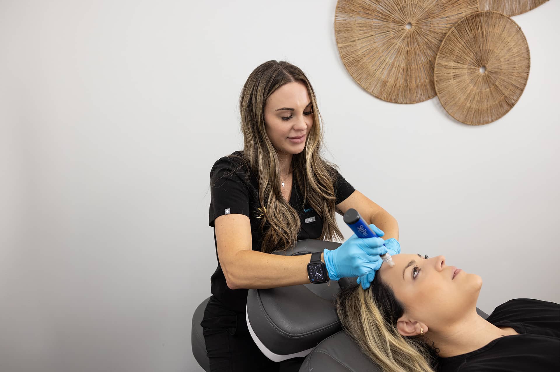 A healthcare professional with long hair and wearing black scrubs and blue gloves is performing a procedure on a patient's forehead with a medical device. The patient, lying back on an examination chair with their head tilted slightly, has light-colored hair. The background has a white wall with round woven decorations.