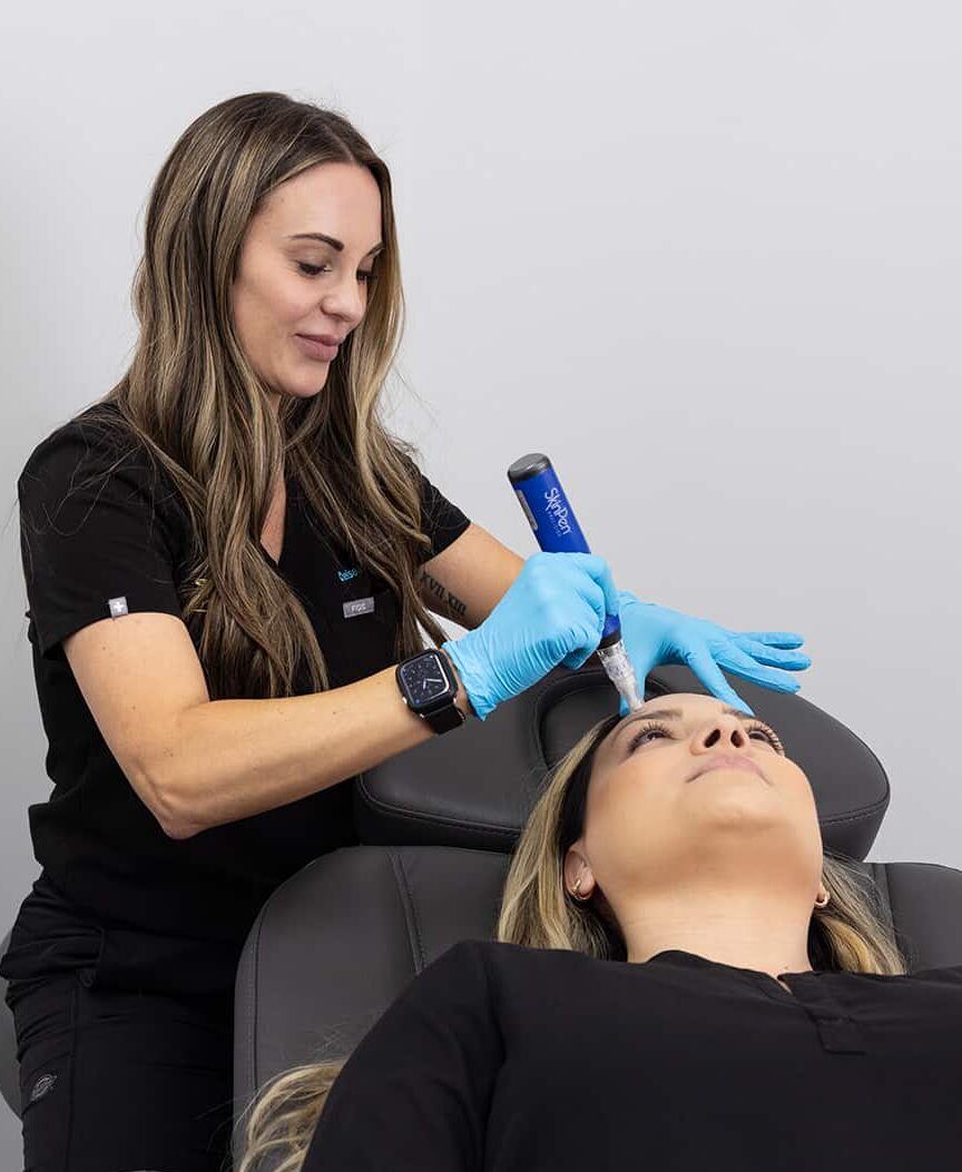 A woman in a black uniform and blue gloves is using a medical device on the forehead of a reclining patient in a clinical setting. There is a tray beside them that holds a brown bottle and a white box. The background is plain white.