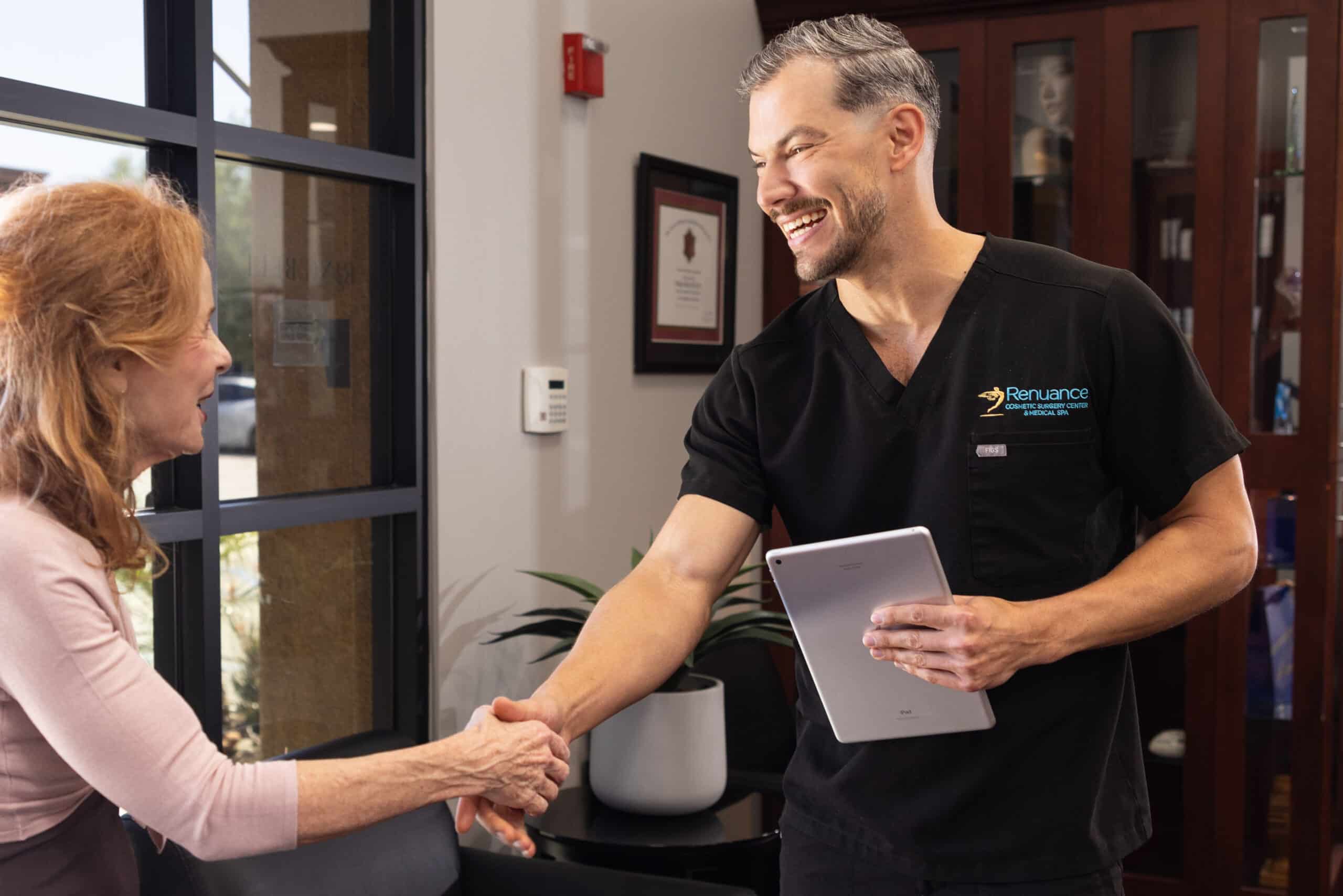 A man wearing black scrubs and holding a tablet is shaking hands with an older woman with red hair in a clinic or medical office setting. The man is smiling and appears to be engaged in conversation with the woman. In the background, there is a glass door, some framed certificates, and a plant.