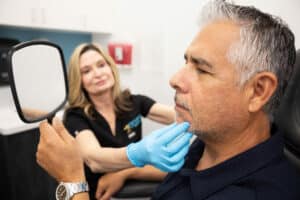 A middle-aged man with gray hair is sitting in a clinic examination room, holding a mirror and looking at his face. A healthcare professional wearing blue gloves is gently touching his chin and smiling, indicating a consultative or examination scenario. The room has white cabinets and a visible red emergency device on the wall.