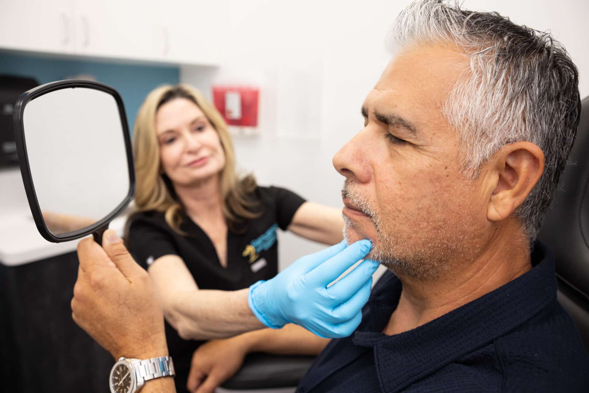 A middle-aged man with gray hair is sitting in a clinic examination room, holding a mirror and looking at his face. A healthcare professional wearing blue gloves is gently touching his chin and smiling, indicating a consultative or examination scenario. The room has white cabinets and a visible red emergency device on the wall.