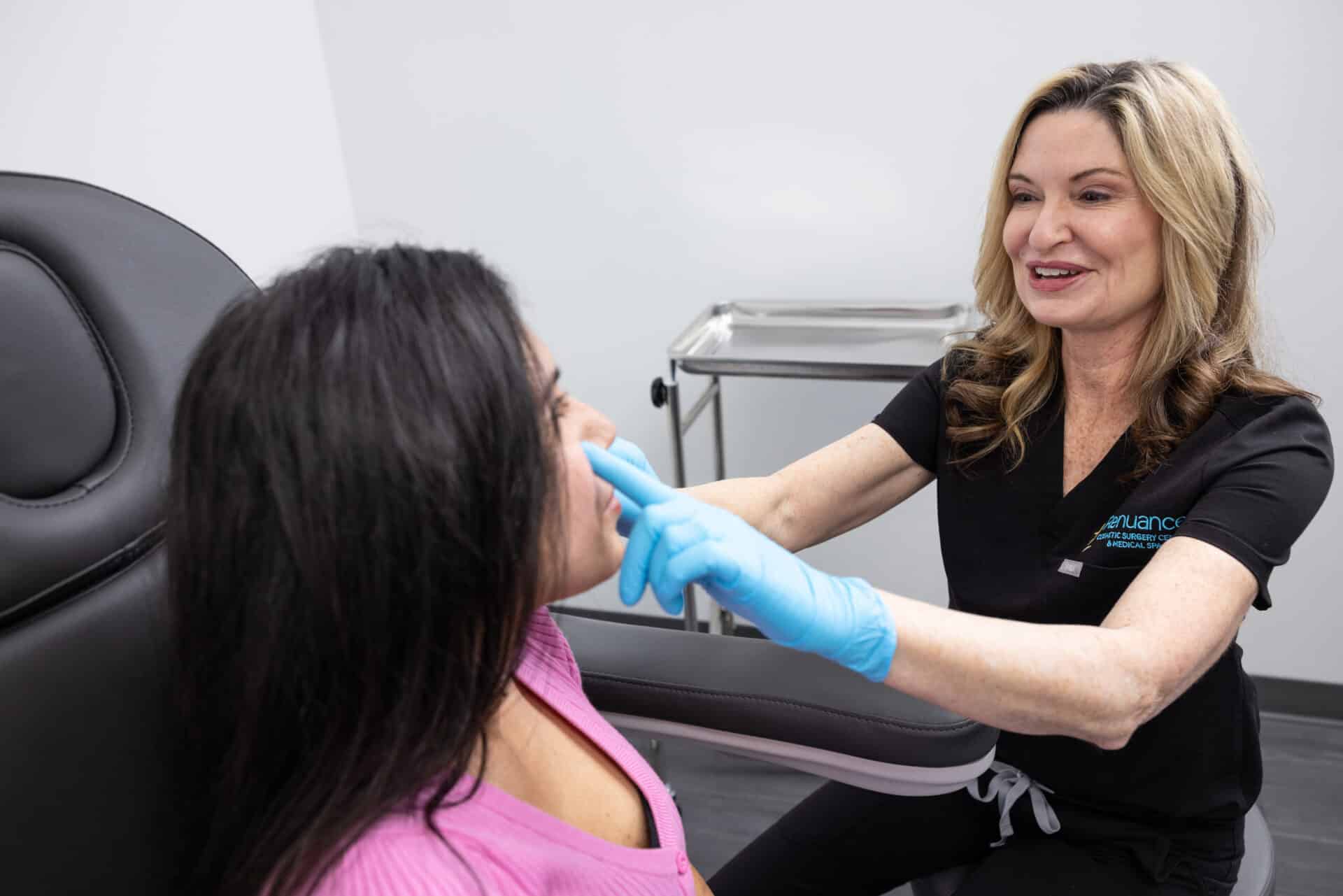A medical professional wearing a black uniform and blue gloves is examining the face of a patient with long dark hair who is sitting in a medical chair. The patient is wearing a pink top. In the background, there is a stainless steel medical tray and a white wall.