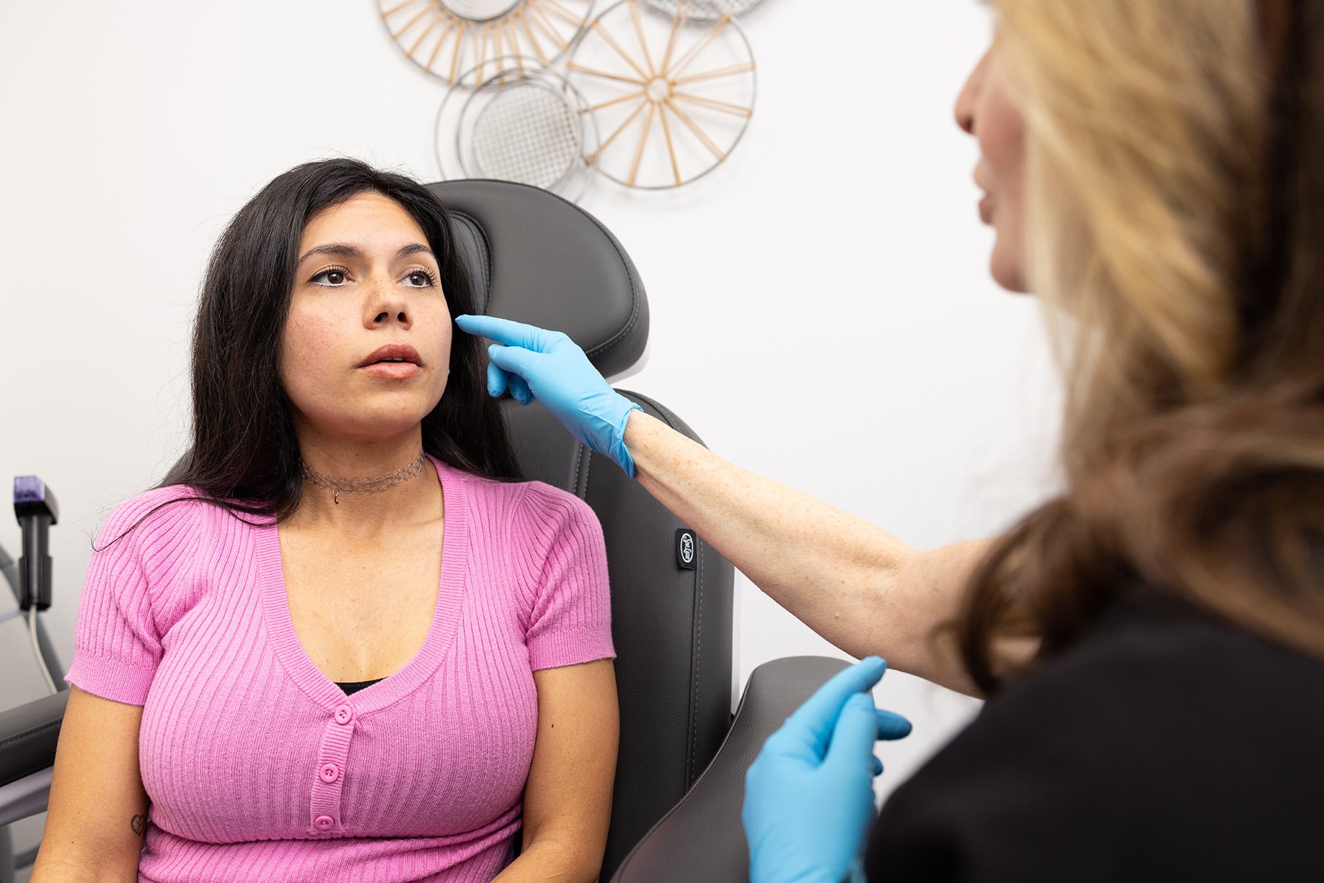 A woman with long dark hair, dressed in a pink ribbed shirt, is sitting on a medical examination chair. She appears to be in a consultation with a healthcare professional, who is wearing blue gloves. The healthcare professional is gently touching the woman's cheek, possibly discussing a medical or cosmetic procedure. The background includes some decorative items on the wall.