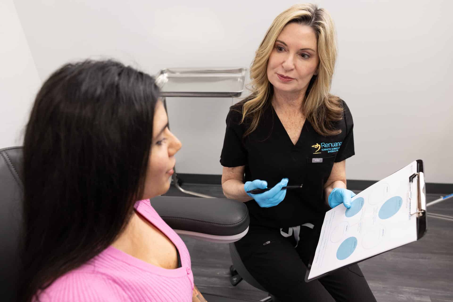 A woman in a black medical uniform and blue gloves is seated while holding a clipboard with charts. She is conversing with another woman who has long dark hair and is wearing a pink sweater. The setting appears to be a medical or consultation room.