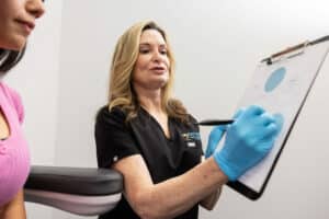 A female healthcare professional wearing black scrubs and blue gloves is holding a clipboard and talking to a patient. The patient, who is visible in the foreground and wearing a pink shirt, is seated with her arm resting on a cushion. The healthcare professional appears to be explaining something written on the clipboard. The setting looks like a medical office.