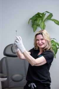 A blonde woman wearing black medical scrubs and white gloves is holding a syringe with a smile in a clinical setting. She stands in front of a chair with a plant in the background.