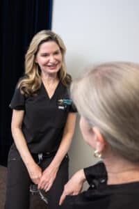 A healthcare professional with long blonde hair, wearing a dark scrub top, is sitting and smiling while engaging in conversation with an older woman whose back is to the camera. The setting appears to be inside a medical office.
